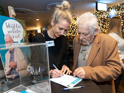 vrouw helpt oudere man met invullen formulier achter een hoge tafel